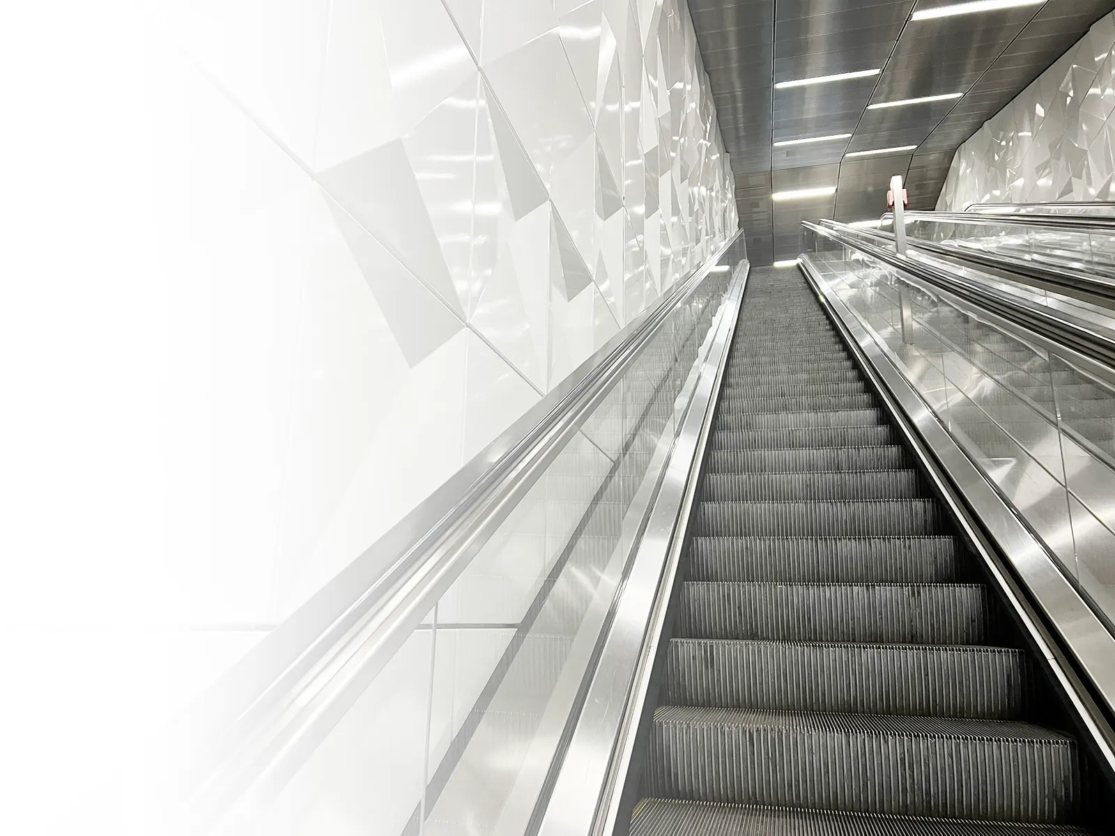 Rolltreppe mit silbernen Handläufen vor weißen Fliesen.